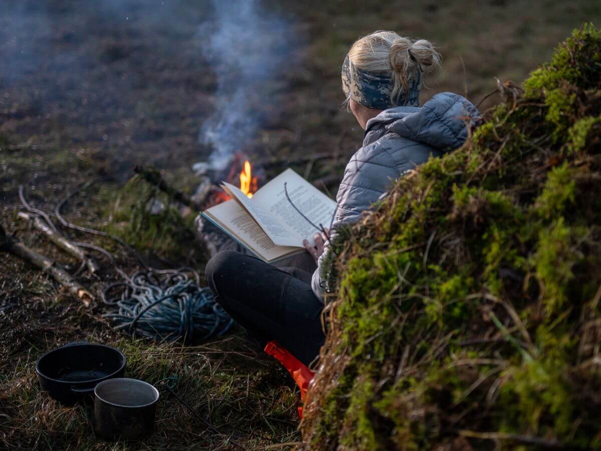 Žena čítajúca knihu v lese pri ohni.