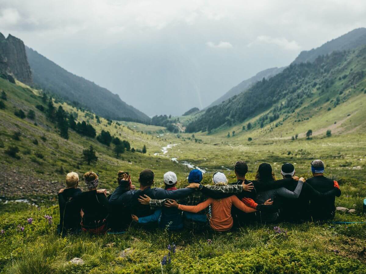 Pohľad zozadu na skupinku ľudí držiacu sa za ramená na túre v prírode.